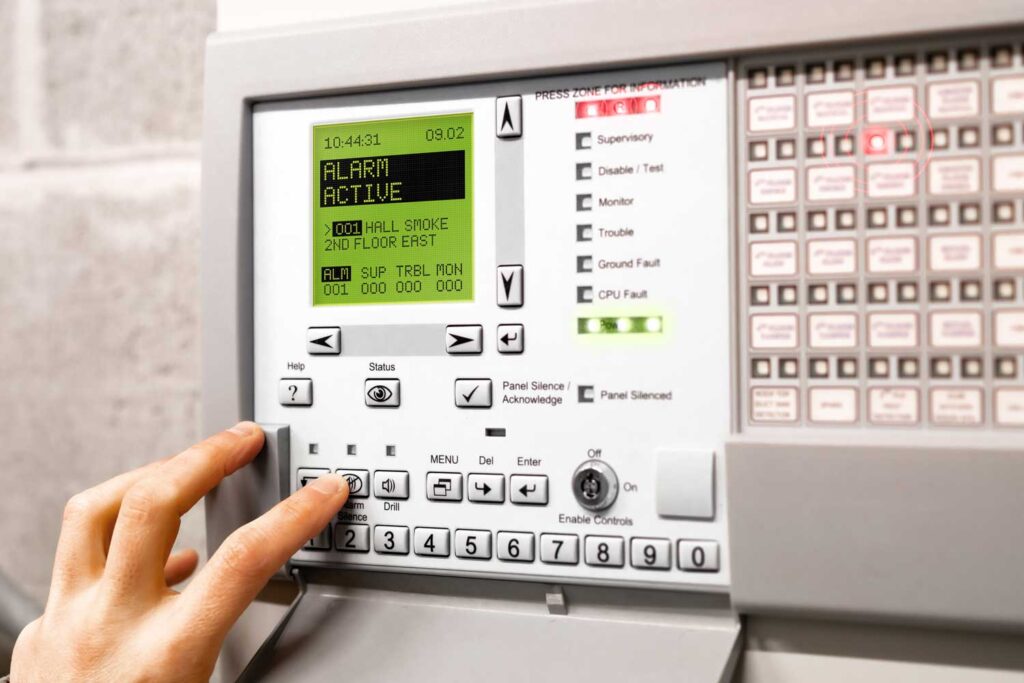 A hand presses a button on a fire alarm control panel displaying an active alarm for smoke detected on the second floor, highlighting the importance of proper fire alarm installation. Various buttons, indicator lights, and a digital screen are visible on the panel.